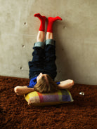 A person lying on a brown shag Finarte Bevi rug against a concrete wall, wearing dark pants, a blue top, and bright red socks, with a colorful Palsta cushion under their head.