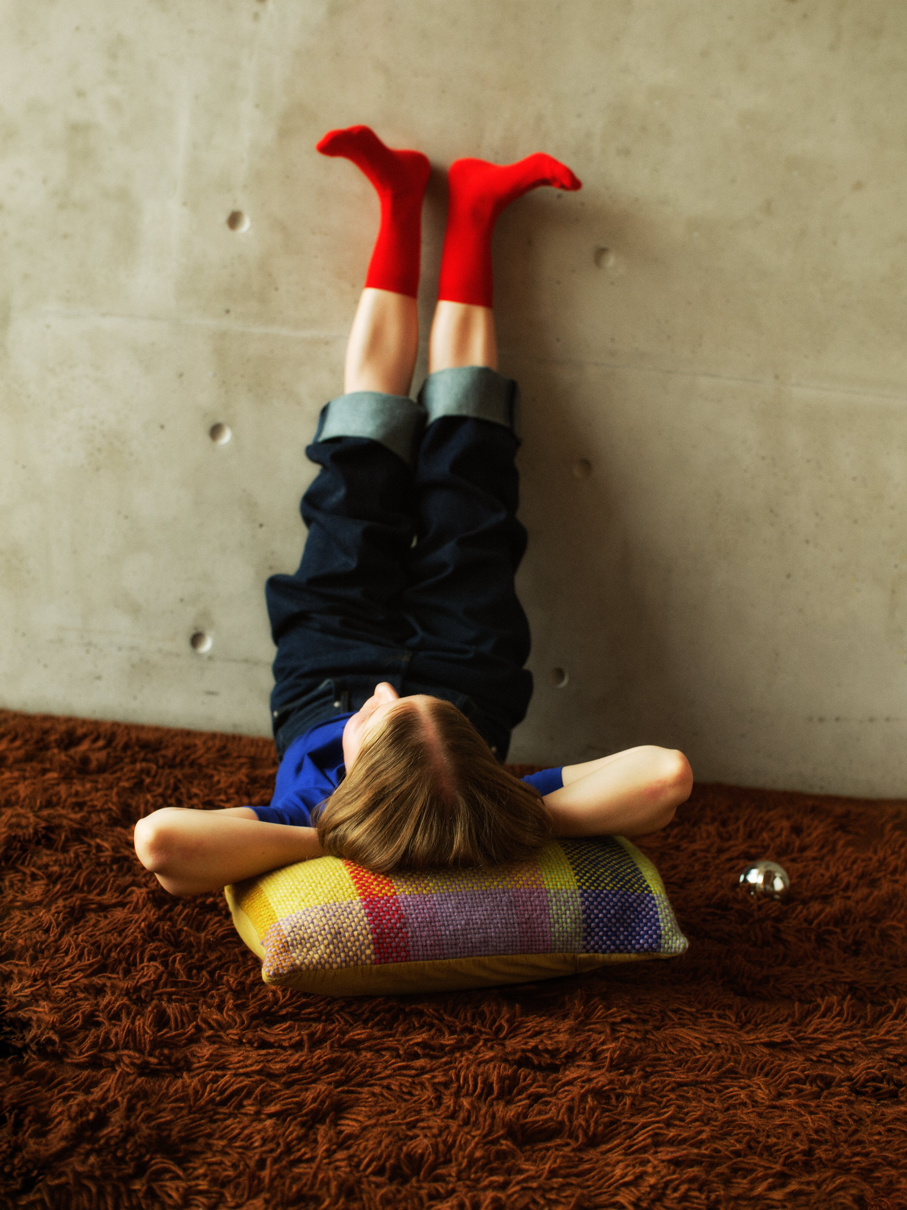 A person lying on a brown shag Finarte Bevi rug against a concrete wall, wearing dark pants, a blue top, and bright red socks, with a colorful Palsta cushion under their head.